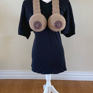 May include: A crocheted brown and tan breast-shaped necklace on a white mannequin wearing a black t-shirt.