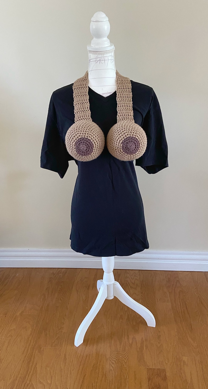 May include: A crocheted brown and tan breast-shaped necklace on a white mannequin wearing a black t-shirt.