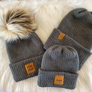 May include: Three grey knitted hats with pom poms. The hats have leather patches with the words "mama", "dada", and "mini" on them.
