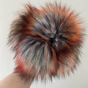 May include: A fluffy faux fur pom pom in shades of black, orange, and grey. The pom pom is made of soft, plush fur and is perfect for adding a touch of whimsy to any hat or accessory.