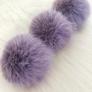 May include: Three fluffy, lavender pom-poms are arranged on a white, textured surface. The pom-poms have a soft, rounded shape and are made of a dense, furry material. The colour is a muted purple with hints of grey.