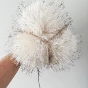 May include: A fluffy, spherical pom-pom crafted from faux fur in a creamy off-white shade, featuring long, wispy strands with dark, charcoal-colored tips that create a textured, multi-dimensional appearance and a soft, voluminous aesthetic.
