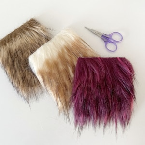 May include: Three pieces of faux fur fabric in different colours: brown, beige, and burgundy. The fabric is cut into squares and laid out on a white surface.