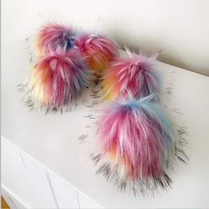Peut inclure: Un ensemble de quatre pompons ronds et duveteux présentant un design en fausse fourrure aux couleurs vives et variées. Chaque pompon affiche un mélange de teintes rose, jaune et bleu avec de longues fibres douces créant une forme sphérique.