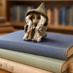 May include: A small ceramic figurine of a seated creature wearing a pointed green hat and long white hair. The figure has a brown face and body, and is positioned on a stack of three books, with a wooden ruler showing 3 inches visible.