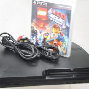 May include: A black PlayStation 3 video game console with a coiled black power cord and a copy of The LEGO Movie Videogame for PS3. The game case features colorful character artwork and the title text clearly displayed on the front cover.