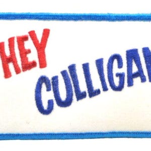 Puede incluir: Un parche rectangular bordado con fondo blanco y borde azul. Presenta una ilustración de línea azul de una persona gritando en un auricular de teléfono, con el texto en rojo y azul "HEY CULLIGAN MAN!" y un pequeño símbolo de marca registrada.