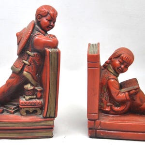 Puede incluir: Par de sujetalibros de cerámica roja que representan a niños con vestimenta tradicional. Un sujetalibros muestra a un niño apoyado en un libro, el otro leyendo. Los sujetalibros tienen detalles dorados y están diseñados para parecer libros.