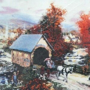 May include: A cross-stitch embroidery depicting a rural landscape with a covered bridge, a horse-drawn carriage, a person fishing by a stream, and vintage cars. The scene features vibrant autumn foliage in shades of orange and red, a blue-roofed bridge, and a small town in the background under a soft, light-colored sky.