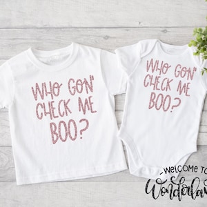 以下が含まれることがあります： ピンクのグリッター文字で「WHO GON' CHECK ME BOO？」と書かれた白いTシャツとベビーボディスーツのセット。お揃いの衣装は、兄弟姉妹や親子での組み合わせを想定しています。このフレーズは遊び心のあるフォントで書かれています。