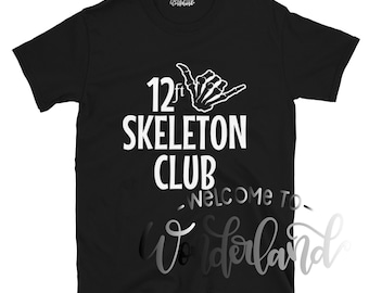 May include: Black t-shirt with white text that reads "12 ft Skeleton Club - Welcome to Wonderland". The text is in a fun, spooky font. A white skeleton hand with a shaka sign is above the text.