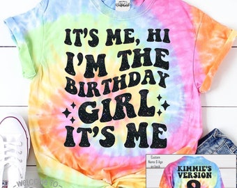 May include: A tie-dye t-shirt with the text "IT'S ME, HI I'M THE BIRTHDAY GIRL IT'S ME" in black glitter lettering. The shirt has a rainbow color scheme and a custom version with the name "KIMMIE'S VERSION" and the number "8" on the back.