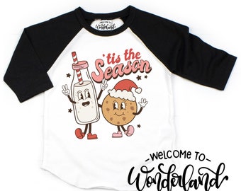 May include: White and black raglan t-shirt featuring a cartoon milk bottle and cookie with smiling faces. The text "'tis the Season" is above the characters. The words "Welcome to Wonderland" are written in script.