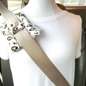 May include: A tan car seatbelt with a leopard print fabric cover. The cover is made of two pieces that are attached to the seatbelt with velcro. The cover is designed to make the seatbelt more comfortable.