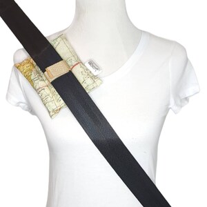 May include: A black seatbelt with a small, rectangular, map-patterned cushion attached to it. The cushion is secured to the seatbelt with a brown fabric strap.
