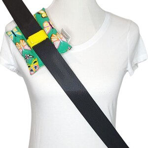 May include: A pair of teal seatbelt covers with a colorful butterfly pattern. The covers are attached to a black seatbelt with a yellow strap. The seatbelt is worn over a white t-shirt.