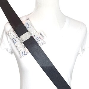 May include: A seat belt cover with a floral pattern in shades of gray, white, and blue. The cover is secured with a silver-colored, sparkly buckle. The seat belt is black.