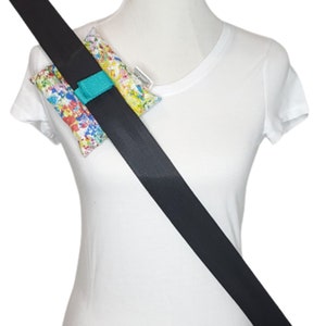 May include: A black seatbelt with a turquoise strap holding a colorful fabric pouch with a white background. The pouch is decorated with a splatter paint design.