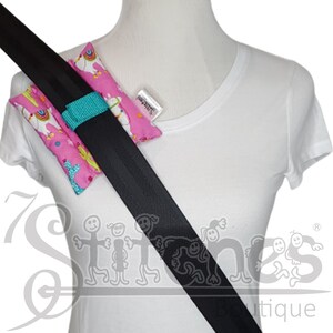 May include: Pink seatbelt cover with a llama and cactus pattern. The cover is secured with a turquoise strap. The seatbelt is black. A white tag is attached to the cover. The cover is designed to provide comfort and protection.
