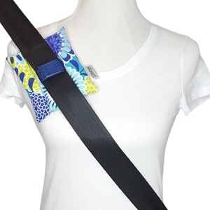May include: A seat belt cover with a blue, yellow, and white floral pattern. The cover is attached to a black seat belt with a blue velcro strap. The seat belt is positioned across a white t-shirt.