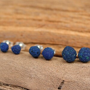 May include: A set of six blue azurite gemstone earrings. The earrings are round and have a rough, natural texture. They are displayed on a wooden surface.