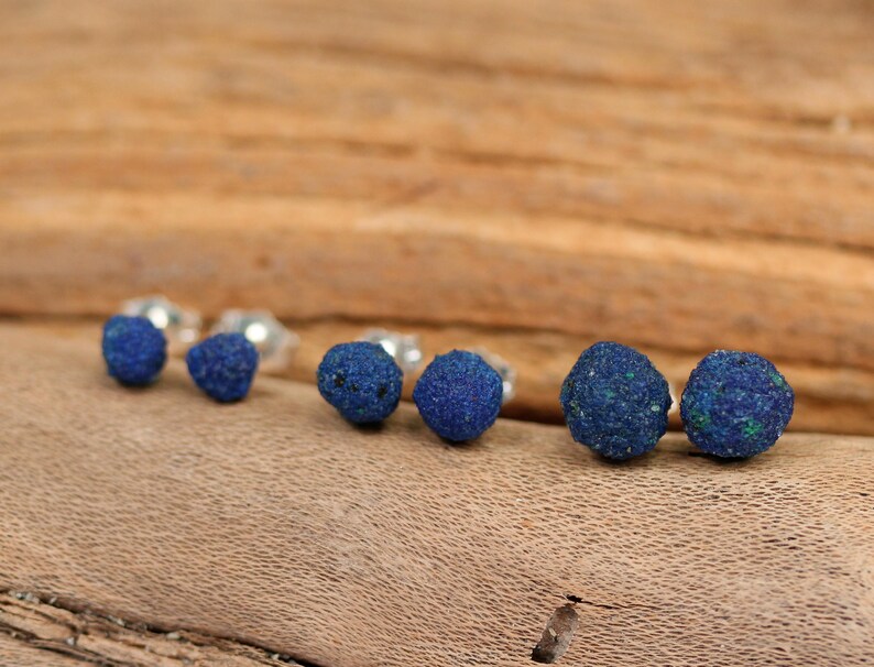 May include: A set of six blue azurite gemstone earrings. The earrings are round and have a rough, natural texture. They are displayed on a wooden surface.