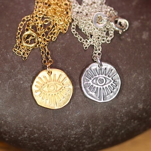 May include: Two necklaces with eye pendants. One necklace has a gold chain and a gold pendant with a detailed eye design. The other necklace has a silver chain and a silver pendant with a detailed eye design.