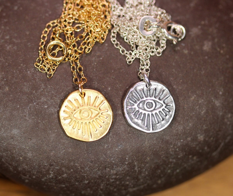 May include: Two necklaces with eye pendants. One necklace has a gold chain and a gold pendant with a detailed eye design. The other necklace has a silver chain and a silver pendant with a detailed eye design.