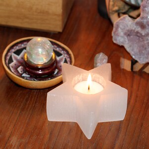 May include: A white star-shaped candle holder with a small tea light candle burning inside. The candle holder is made of selenite crystal and has a rough, textured surface. The candle holder is sitting on a wooden table.