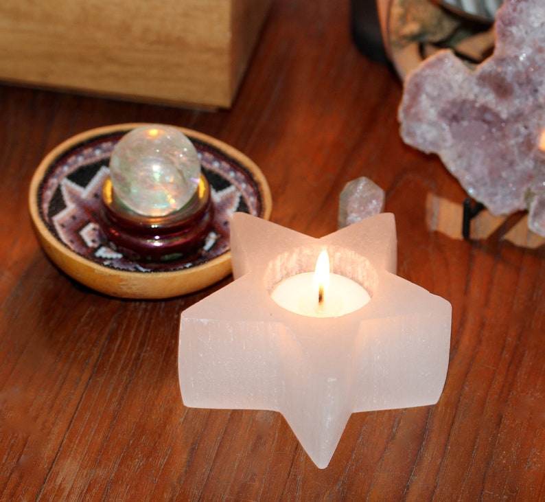 May include: A white star-shaped candle holder with a small tea light candle burning inside. The candle holder is made of selenite crystal and has a rough, textured surface. The candle holder is sitting on a wooden table.