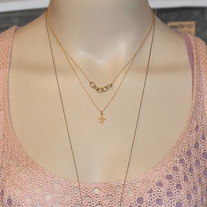 May include: A layered gold necklace with a small pendant in the shape of a bird. The necklace is worn on a mannequin with a pink crocheted top.