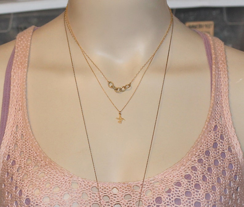 May include: A layered gold necklace with a small pendant in the shape of a bird. The necklace is worn on a mannequin with a pink crocheted top.