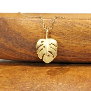 Peut inclure: Un collier en or avec un pendentif d&eacute;licat en forme de feuille de monstera d&eacute;coup&eacute;e. Le collier est pr&eacute;sent&eacute; sur une surface en bois.
