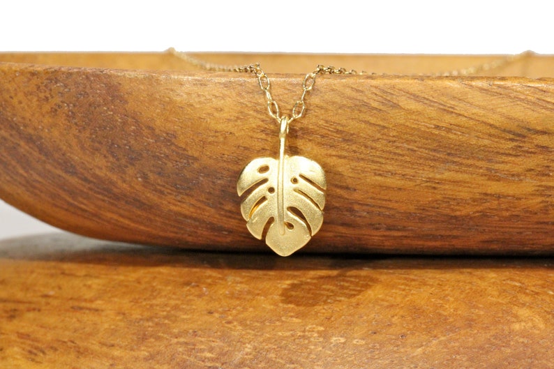 Peut inclure: Un collier en or avec un pendentif d&eacute;licat en forme de feuille de monstera d&eacute;coup&eacute;e. Le collier est pr&eacute;sent&eacute; sur une surface en bois.