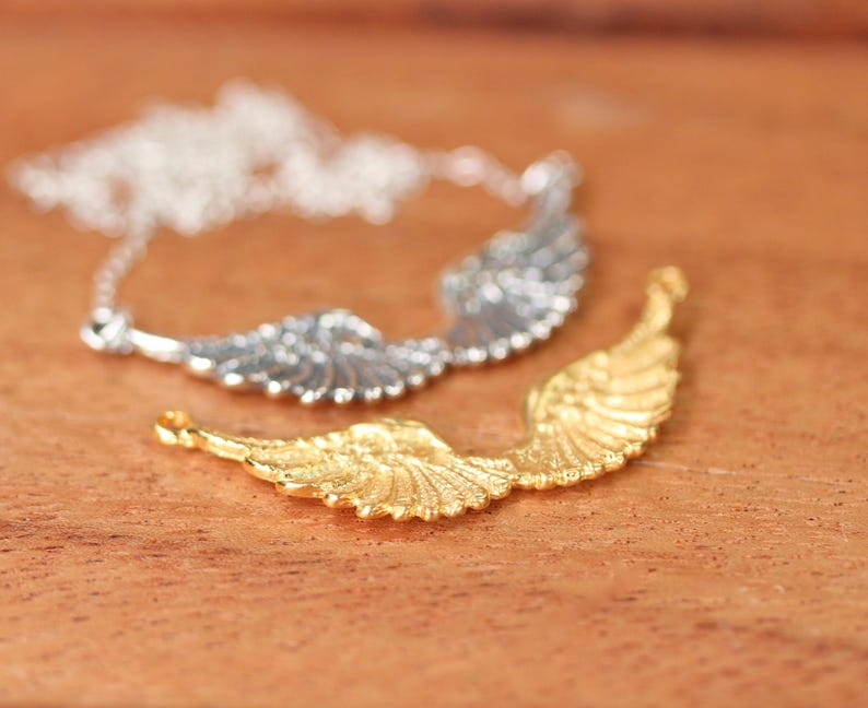 Puede incluir: Dos abalorios con forma de alas de &aacute;ngel, uno plateado y otro dorado, sobre una superficie de madera. El abalorio dorado est&aacute; enfocado y el plateado est&aacute; desenfocado.