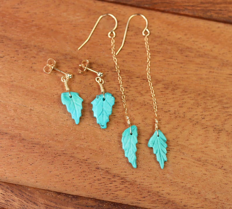 May include: A pair of turquoise leaf earrings with gold chain and hook closures. The earrings are on a wooden surface.