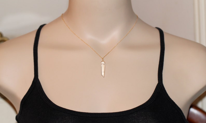May include: A gold chain necklace with a clear quartz crystal pendant. The pendant is a long, thin, pointed shape.