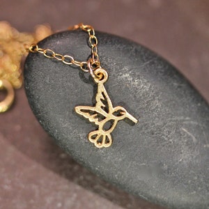 May include: A gold chain necklace with a delicate hummingbird pendant. The hummingbird is cut out of metal and has a detailed design.