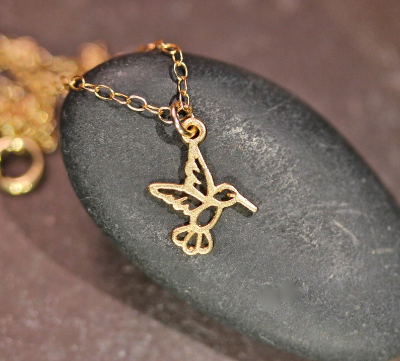 May include: A gold chain necklace with a delicate hummingbird pendant. The hummingbird is cut out of metal and has a detailed design.