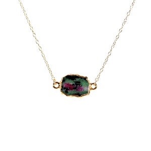 May include: A gold-colored necklace featuring a raw, rectangular gemstone pendant. The pendant has a green and pink color scheme, with a gold-colored bezel. The necklace chain is delicate and gold-colored.