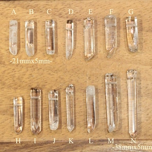 May include: A set of 14 clear quartz crystal points, labeled A through N, arranged in two rows on a wooden surface. The crystals are approximately 21 mm x 5 mm and 38 mm x 5 mm in size.