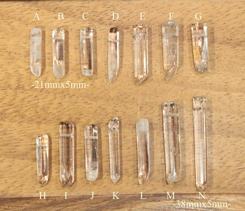 May include: A set of 14 clear quartz crystal points, labeled A through N, arranged in two rows on a wooden surface. The crystals are approximately 21 mm x 5 mm and 38 mm x 5 mm in size.