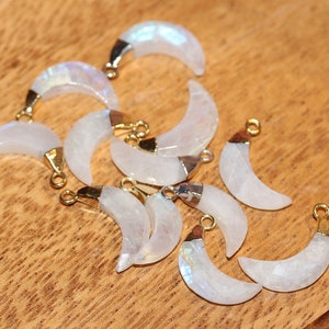 May include: A collection of eight white moonstone crescent charms with gold accents. The charms are arranged on a wooden surface.