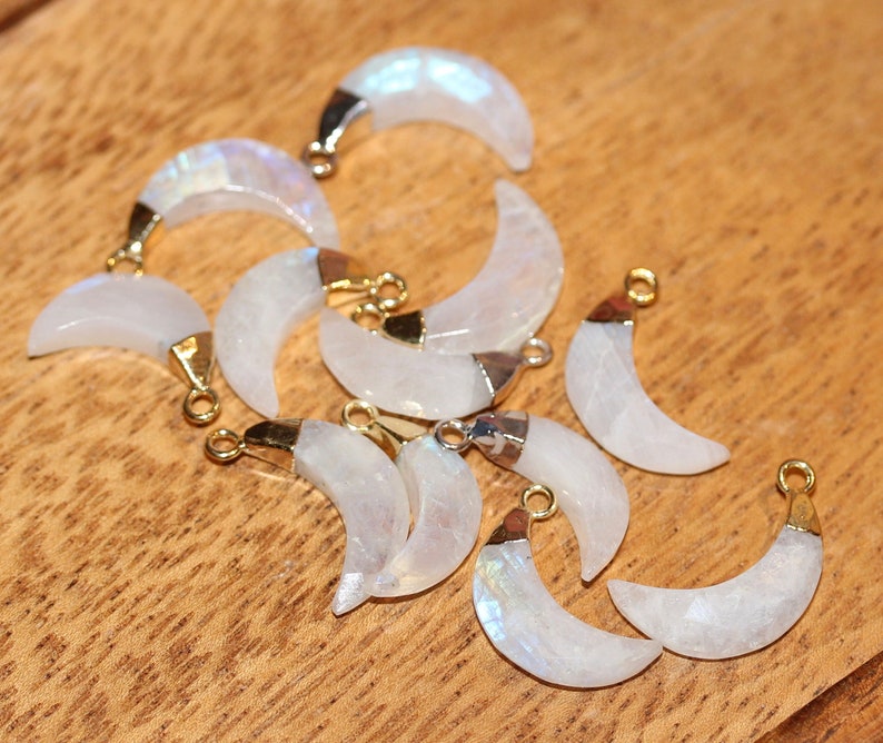 May include: A collection of eight white moonstone crescent charms with gold accents. The charms are arranged on a wooden surface.