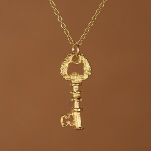 Puede incluir: Un collar con un colgante de llave de oro con un dise&ntilde;o ornamentado detallado. La llave tiene un gran lazo decorativo en la parte superior y un eje largo y delgado.