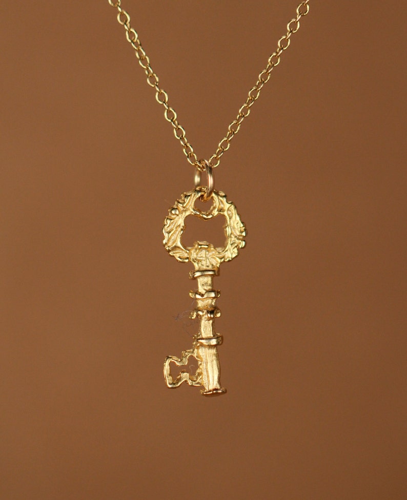Puede incluir: Un collar con un colgante de llave de oro con un dise&ntilde;o ornamentado detallado. La llave tiene un gran lazo decorativo en la parte superior y un eje largo y delgado.