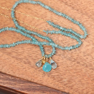 May include: A delicate necklace made with light blue gemstone beads and gold accents. The necklace features a cluster of three gemstone drops, including a larger, round, light blue gemstone.