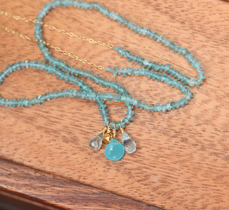 May include: A delicate necklace made with light blue gemstone beads and gold accents. The necklace features a cluster of three gemstone drops, including a larger, round, light blue gemstone.