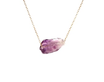 May include: A delicate gold chain necklace with a raw amethyst crystal pendant. The crystal is a deep purple color and has a natural, irregular shape.