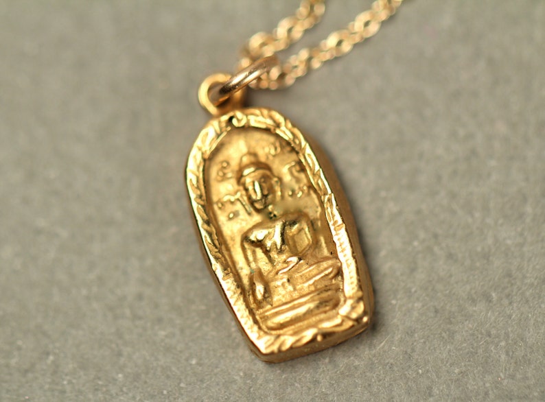 May include: Gold pendant necklace featuring a detailed image of a seated Buddha figure. The pendant is oval shaped and has a decorative border.